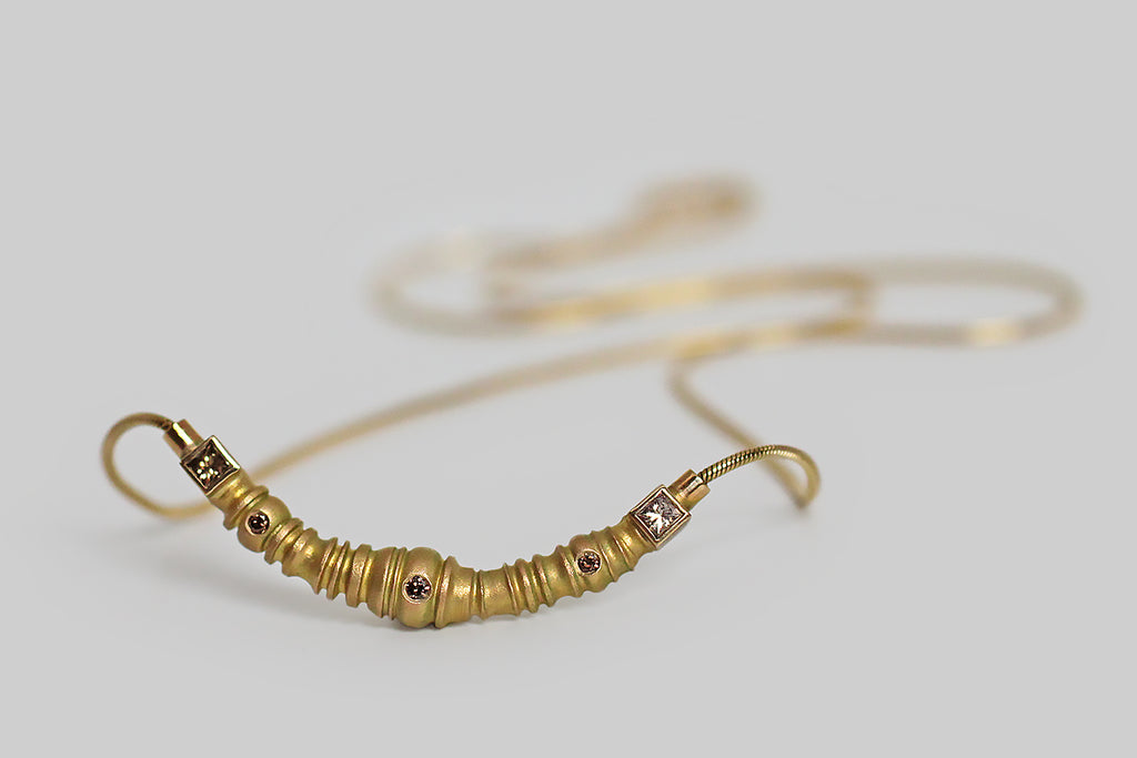 A compelling 1990s necklace, modeled in 14k yellow gold, whose centerpiece is weighty, crescent-shaped, and studded with three flush-set diamonds. This crescent is matte-finished and highly textural; its segmented form is both organized and organic-seeming, with ridges, collars, and nodules that come together to insinuate some kind of biomech— a mechanical caterpillar, or a relic from an HR Geiger illustration. 