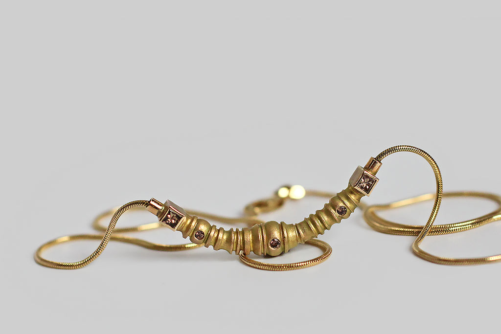 A compelling 1990s necklace, modeled in 14k yellow gold, whose centerpiece is weighty, crescent-shaped, and studded with three flush-set diamonds. This crescent is matte-finished and highly textural; its segmented form is both organized and organic-seeming, with ridges, collars, and nodules that come together to insinuate some kind of biomech— a mechanical caterpillar, or a relic from an HR Geiger illustration. 