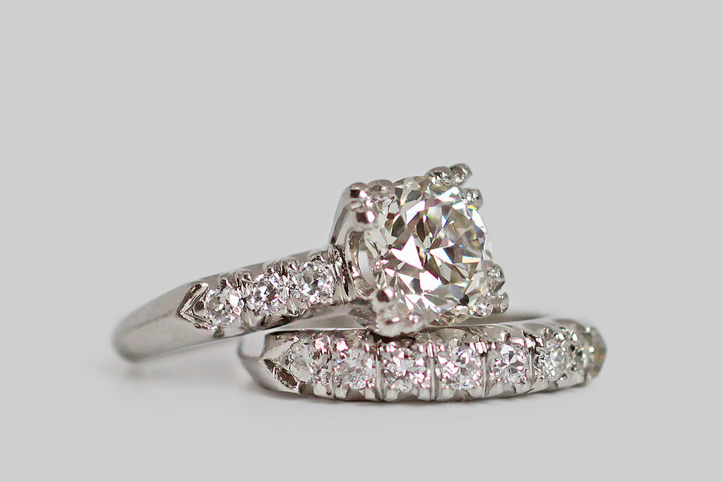 An elegant, 1940s wedding set, modeled in platinum, whose engagement ring features a soulful, 2 carat, old European cut diamond. This lively, hand-cut darling is held in aloft in four, three-lobed prongs and is flanked by trios of smaller diamonds— these supporters are held in fishtail settings across the ring's pitched shoulders. The engagement ring's substantial, knife-edge shank tapers toward the base.