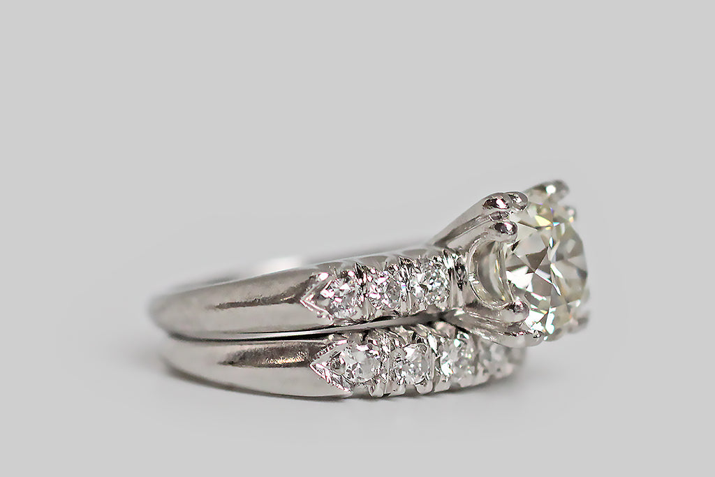 An elegant, 1940s wedding set, modeled in platinum, whose engagement ring features a soulful, 2 carat, old European cut diamond. This lively, hand-cut darling is held in aloft in four, three-lobed prongs and is flanked by trios of smaller diamonds— these supporters are held in fishtail settings across the ring's pitched shoulders. The engagement ring's substantial, knife-edge shank tapers toward the base.