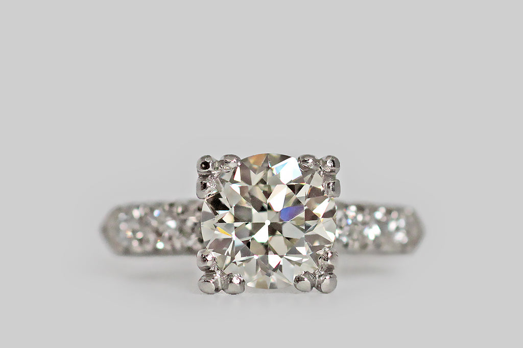 An elegant, 1940s wedding set, modeled in platinum, whose engagement ring features a soulful, 2 carat, old European cut diamond. This lively, hand-cut darling is held in aloft in four, three-lobed prongs and is flanked by trios of smaller diamonds— these supporters are held in fishtail settings across the ring's pitched shoulders. The engagement ring's substantial, knife-edge shank tapers toward the base.