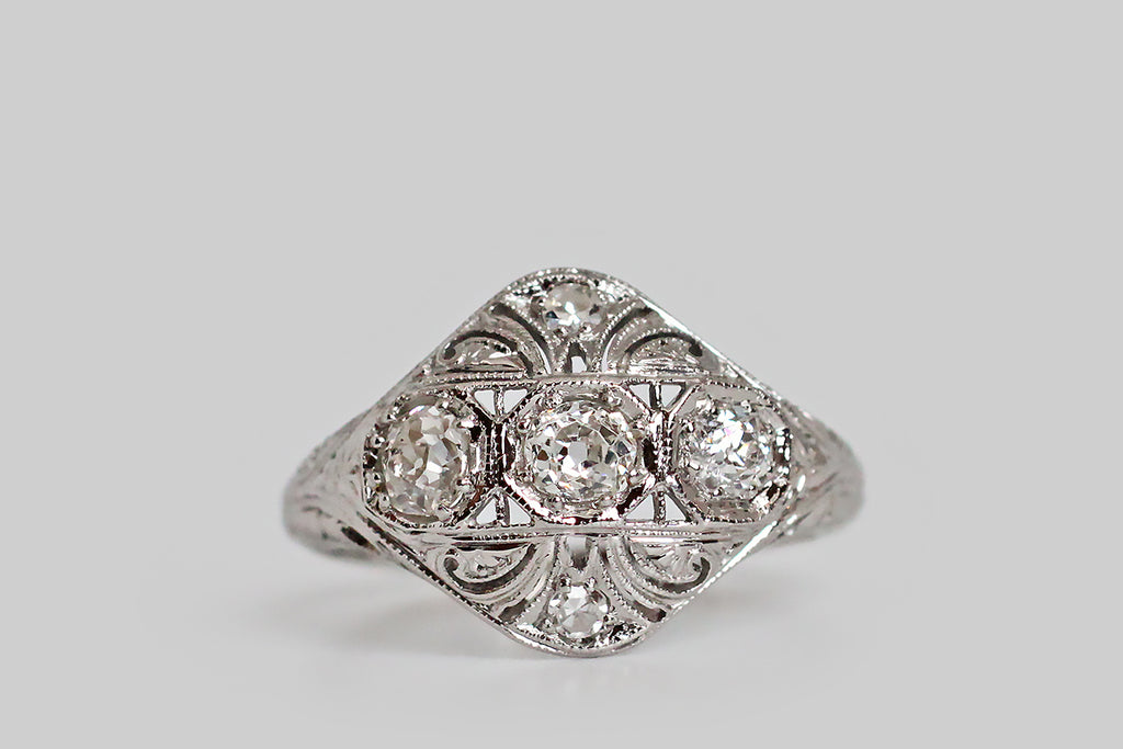 An Art Deco era, diamond-set, panel ring, with a softly-bowed, diamond-shaped head. This ornate old darling has a delicately pierced face, that is decorated with a series of plumes, or flourishes, which seem to curl out from its center. The ring's three primary diamonds are charming old mine cuts; they are bead-set horizontally, into hex-shaped seats, across the center of the ring face. 