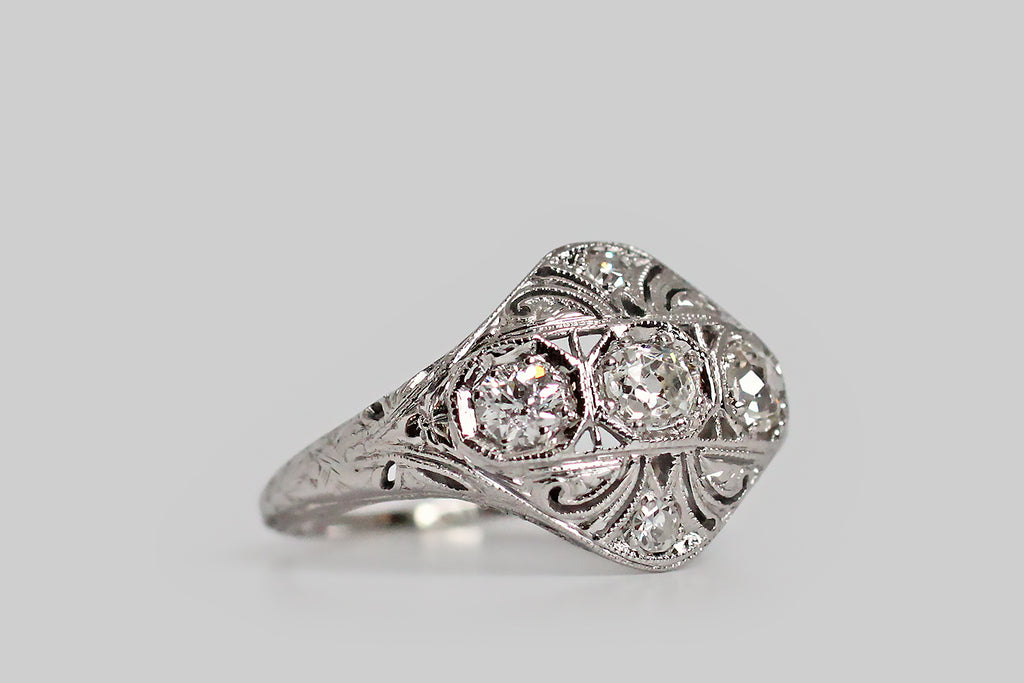 An Art Deco era, diamond-set, panel ring, with a softly-bowed, diamond-shaped head. This ornate old darling has a delicately pierced face, that is decorated with a series of plumes, or flourishes, which seem to curl out from its center. The ring's three primary diamonds are charming old mine cuts; they are bead-set horizontally, into hex-shaped seats, across the center of the ring face. 