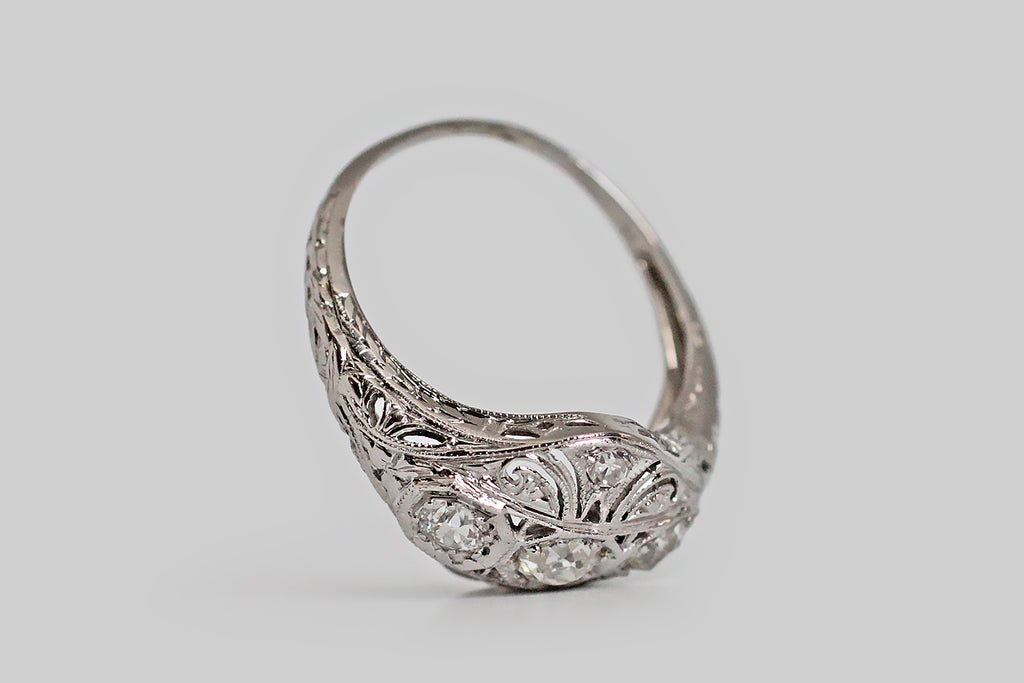 An Art Deco era, diamond-set, panel ring, with a softly-bowed, diamond-shaped head. This ornate old darling has a delicately pierced face, that is decorated with a series of plumes, or flourishes, which seem to curl out from its center. The ring's three primary diamonds are charming old mine cuts; they are bead-set horizontally, into hex-shaped seats, across the center of the ring face. 
