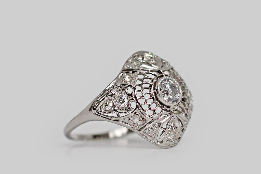 An Art Deco era, diamond cluster ring, modeled in platinum, with a gently-bowed, diamond-shaped head. This ring is visually delicate— its central decor is a fine, hand-pierced series of scalloped cells that gives the impression of a spiderweb, or perhaps of layered feathers. Fleur de lis shaped seats are posted at the ring's cardinal points (NSEW); each of these is set with single cut diamonds diamonds, and a handful of smaller, single cut supporters sparkle around the perimeter of the ring face.