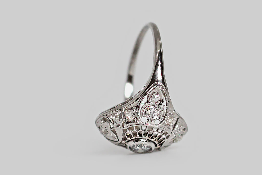An Art Deco era, diamond cluster ring, modeled in platinum, with a gently-bowed, diamond-shaped head. This ring is visually delicate— its central decor is a fine, hand-pierced series of scalloped cells that gives the impression of a spiderweb, or perhaps of layered feathers. Fleur de lis shaped seats are posted at the ring's cardinal points (NSEW); each of these is set with single cut diamonds diamonds, and a handful of smaller, single cut supporters sparkle around the perimeter of the ring face.