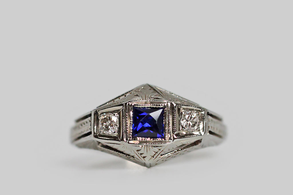 A stylized, Art Deco engagement ring, modeled in 18k white gold, whose trio of gems are a French cut, created, blue sapphire, and a sparkling pair of transitional-cut, white diamonds. These gems are oriented in a tidy, horizontal row, where they are held in square seats— each of these seats is finished with a fine milgrain edge. This ring has a unique, dimensional quality, that gives it an aerodynamic feel