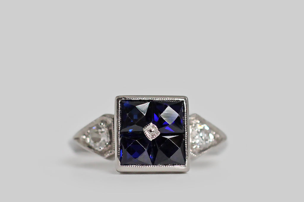 Poor Mouchette | Curated Antique Jewelry, Vintage Jewelry & Engagement Rings | Portland, Oregon | A charming Art Deco era ring, modeled in palladium, whose four, central, French cut sapphires come together to suggest an orange blossom. These deep-blue, created sapphires are set calibre-style (edge to edge) inside their large, square bezel, and a small, milgrained, palladium bead pins them, at center. The ring's square head is flanked by kite-shaped shoulders, each holding a chunky old mine cut diamond.