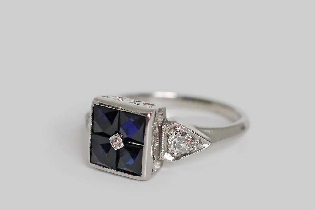 Poor Mouchette | Curated Antique Jewelry, Vintage Jewelry & Engagement Rings | Portland, Oregon | A charming Art Deco era ring, modeled in palladium, whose four, central, French cut sapphires come together to suggest an orange blossom. These deep-blue, created sapphires are set calibre-style (edge to edge) inside their large, square bezel, and a small, milgrained, palladium bead pins them, at center. The ring's square head is flanked by kite-shaped shoulders, each holding a chunky old mine cut diamond.