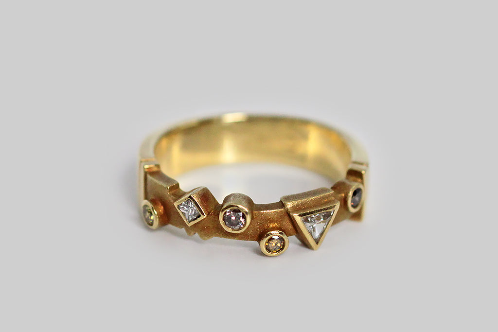 A whimsical 18k gold band, set with an array of fancy colored diamonds, in various shapes. Among these natural, bezel set stones is a cognac round brilliant diamond, two round brilliant canary diamonds, a silver colored round brilliant diamond, and two white diamonds (a triangle and a diamond shape). This playful geometric arrangement sits atop the ring's cutaway face, which has been textured for contrast and features a couple of shapely notches that I especially love. 