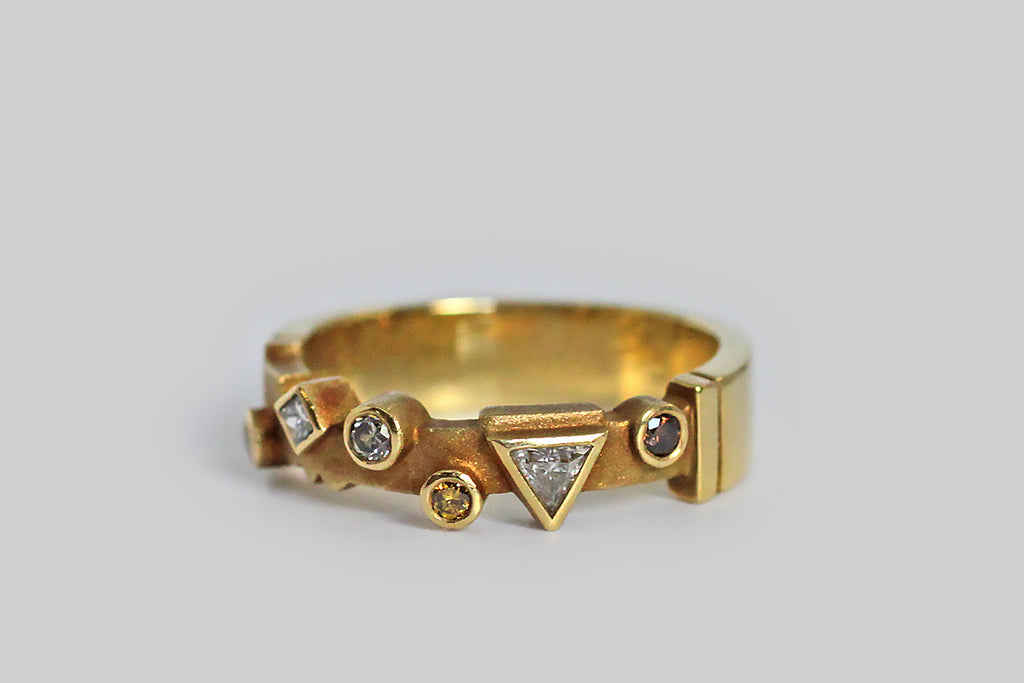 A whimsical 18k gold band, set with an array of fancy colored diamonds, in various shapes. Among these natural, bezel set stones is a cognac round brilliant diamond, two round brilliant canary diamonds, a silver colored round brilliant diamond, and two white diamonds (a triangle and a diamond shape). This playful geometric arrangement sits atop the ring's cutaway face, which has been textured for contrast and features a couple of shapely notches that I especially love.