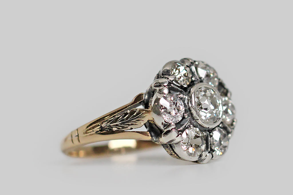 A soulful, Victorian-era, cluster ring, modeled in silver and 14k yellow gold, whose seven OMC diamonds come together in the form of a flower. The chunky, old mine cut diamonds that make up the petals of this flower are set in silver collects, which are reinforced with trios of petal-like prongs— ridge-like silver spaces also "compartmentalize" these diamonds. The ring's central .30 carat diamond is held in a full bezel. The ring's tapering, half-round shank is modeled in 14k yellow gold