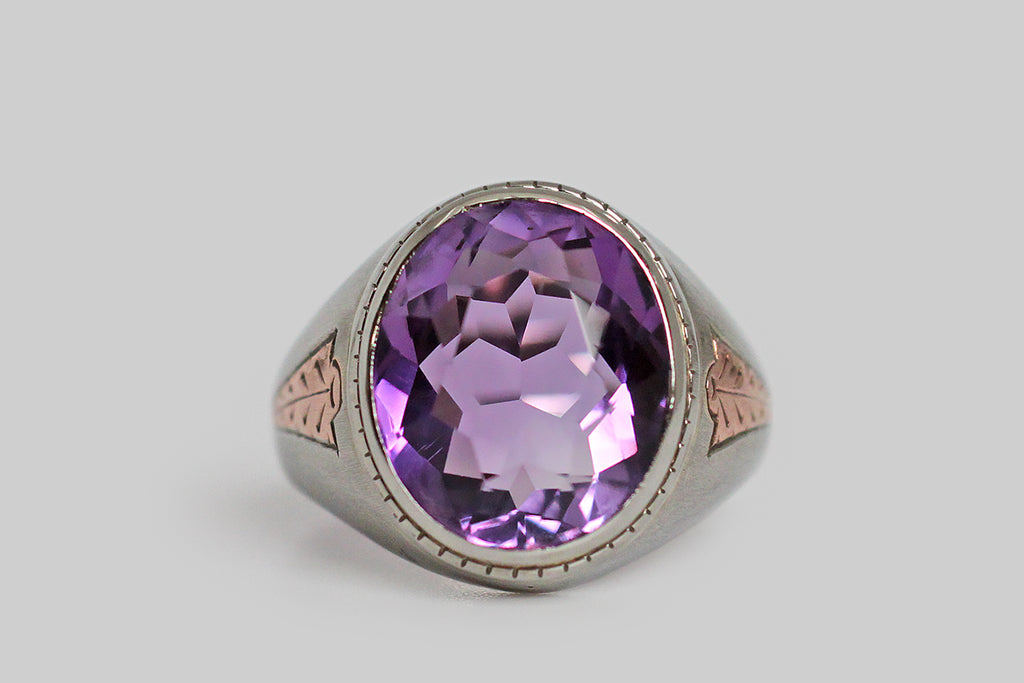 A dandy, streamlined, 1940s ring, modeled in 10k white gold, whose shoulders feature a charming, engraved, rose gold detail. This hand-engraved motif calls to mind an arrowhead, or the tip of a spear— it gives the impression of speed. This ring features a big, beautiful, faceted, amethyst (approx 6.4 carats). The highly-saturated, violet-purple gem is held in a smooth bezel; small hashes decorate the bezel's base. 