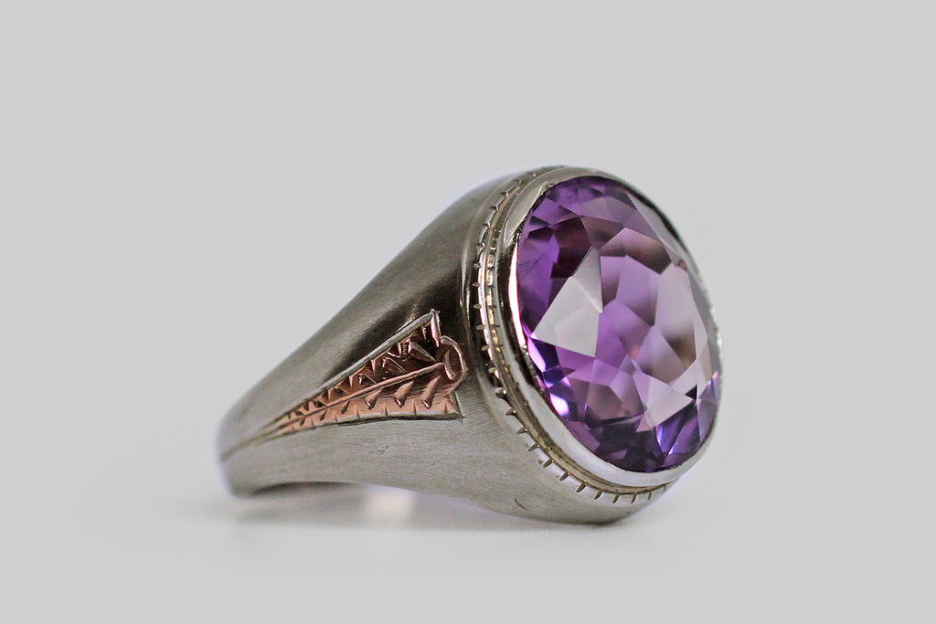 A dandy, streamlined, 1940s ring, modeled in 10k white gold, whose shoulders feature a charming, engraved, rose gold detail. This hand-engraved motif calls to mind an arrowhead, or the tip of a spear— it gives the impression of speed. This ring features a big, beautiful, faceted, amethyst (approx 6.4 carats). The highly-saturated, violet-purple gem is held in a smooth bezel; small hashes decorate the bezel's base. 