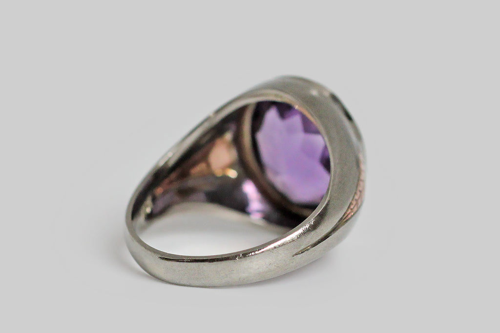 A dandy, streamlined, 1940s ring, modeled in 10k white gold, whose shoulders feature a charming, engraved, rose gold detail. This hand-engraved motif calls to mind an arrowhead, or the tip of a spear— it gives the impression of speed. This ring features a big, beautiful, faceted, amethyst (approx 6.4 carats). The highly-saturated, violet-purple gem is held in a smooth bezel; small hashes decorate the bezel's base. 