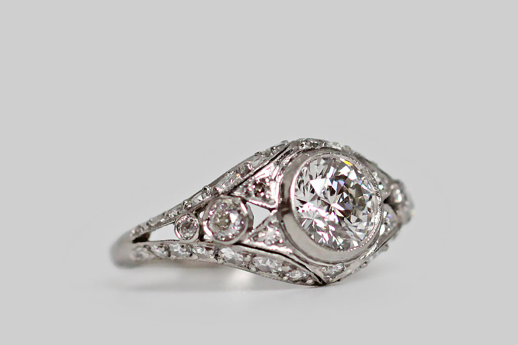 A striking Art Deco era engagement ring, modeled in platinum, featuring a 1.09 carat transitional cut diamond. This lovely central gem (F/SI1) rests in a slim milgrain bezel. It is supported by a bevy of smaller diamonds (both early modern brilliant cuts, and old mine cuts), that fill up both sides of the ring's ornate gallery, and decorate its open-work face. We especially love the diamond-set, cutaway, forked-pennant decor that flanks the primary diamond, like a pair of prim little collars.