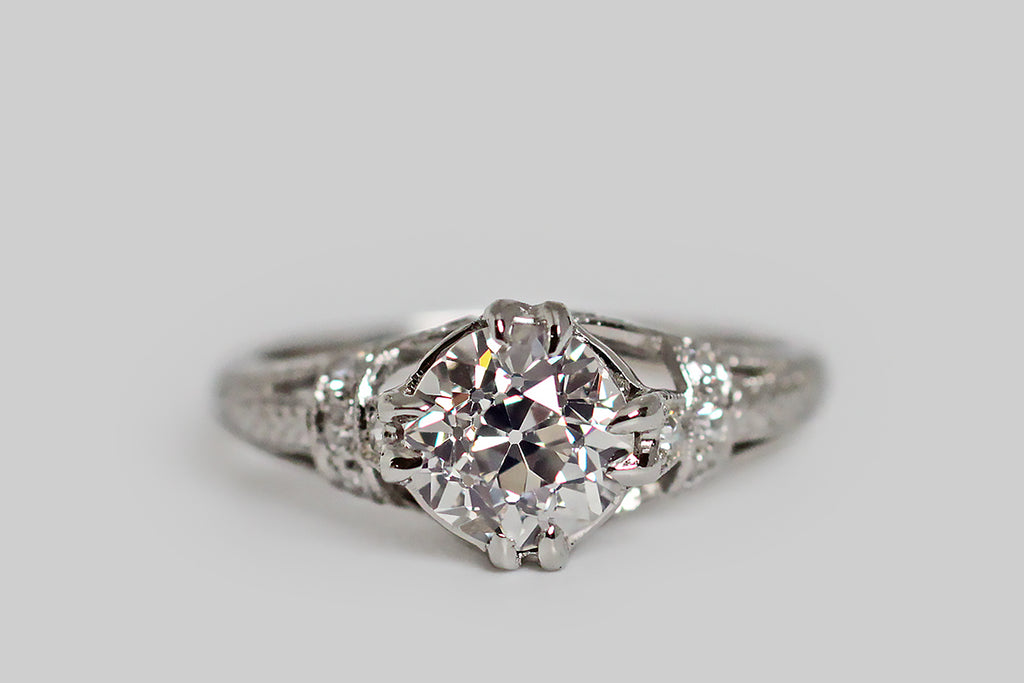 An elegant, Art Deco era engagement ring, modeled in platinum, whose primary gem is a 1.31 carat old mine cut diamond. This lively, central diamond (K-L/SI2) is held in four sets of double prongs. It is supported by a bevy of single cut diamonds, that add sparkle to the ring's intricate shoulders and gallery. This modified cathedral setting features split shoulders that are decoratively bound with diamond-set garters