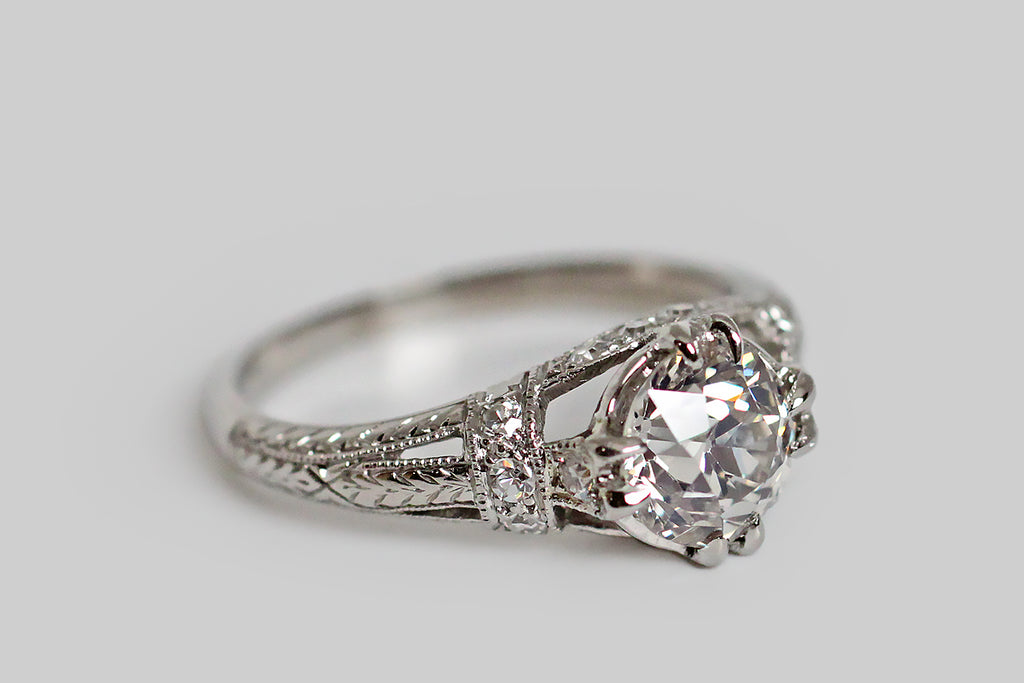 An elegant, Art Deco era engagement ring, modeled in platinum, whose primary gem is a 1.31 carat old mine cut diamond. This lively, central diamond (K-L/SI2) is held in four sets of double prongs. It is supported by a bevy of single cut diamonds, that add sparkle to the ring's intricate shoulders and gallery. This modified cathedral setting features split shoulders that are decoratively bound with diamond-set garters