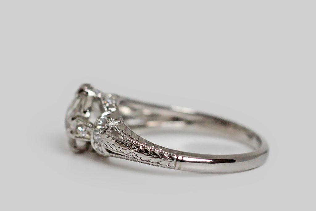 An elegant, Art Deco era engagement ring, modeled in platinum, whose primary gem is a 1.31 carat old mine cut diamond. This lively, central diamond (K-L/SI2) is held in four sets of double prongs. It is supported by a bevy of single cut diamonds, that add sparkle to the ring's intricate shoulders and gallery. This modified cathedral setting features split shoulders that are decoratively bound with diamond-set garters