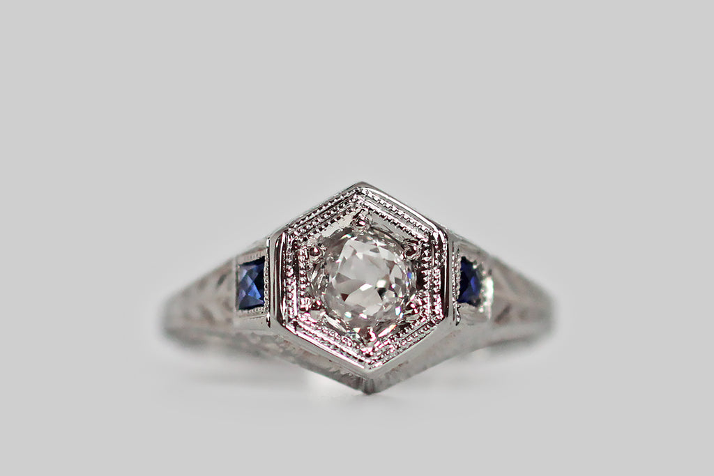 A really lovely 1920s engagement ring, modeled in 18k white gold, whose central gem is a lively, half-carat, old mine cut diamond, which is bead set into the ring's hex-shaped face. This Art Deco era ring is decorated, all-over, with intricate hand-engraving— the shoulders and sides of the ring feature a traditional wheat pattern, while the inter-cardinal points of the ring head are each decorated with an eight-pointed star. 