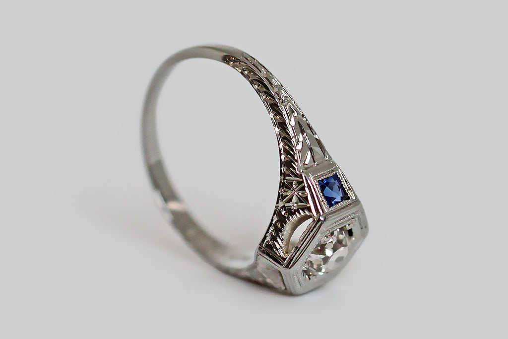 A really lovely 1920s engagement ring, modeled in 18k white gold, whose central gem is a lively, half-carat, old mine cut diamond, which is bead set into the ring's hex-shaped face. This Art Deco era ring is decorated, all-over, with intricate hand-engraving— the shoulders and sides of the ring feature a traditional wheat pattern, while the inter-cardinal points of the ring head are each decorated with an eight-pointed star. 
