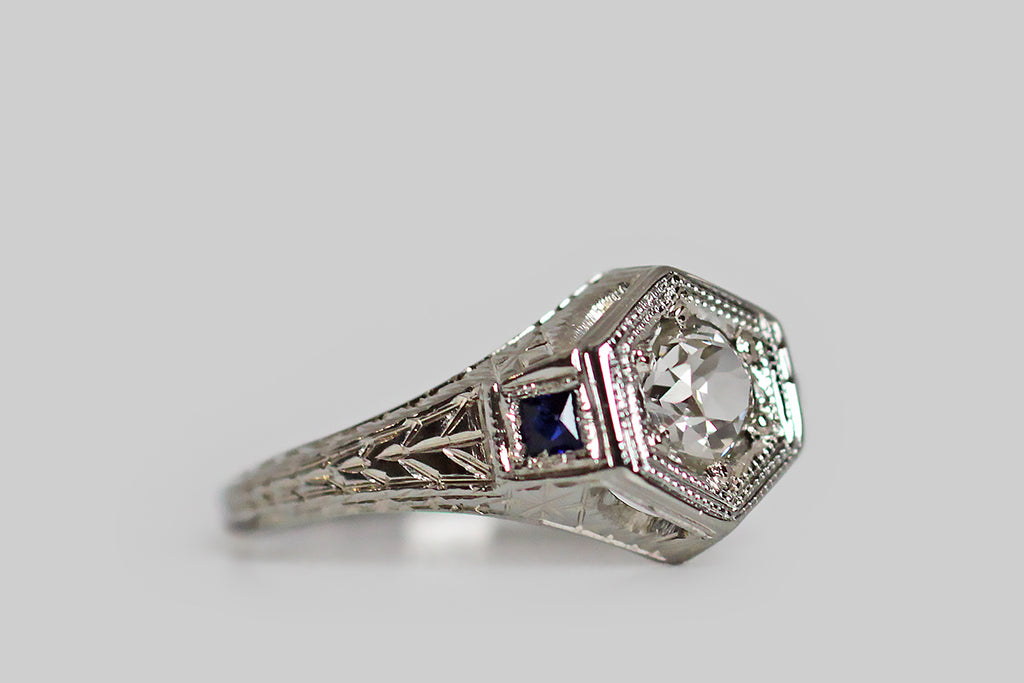 A really lovely 1920s engagement ring, modeled in 18k white gold, whose central gem is a lively, half-carat, old mine cut diamond, which is bead set into the ring's hex-shaped face. This Art Deco era ring is decorated, all-over, with intricate hand-engraving— the shoulders and sides of the ring feature a traditional wheat pattern, while the inter-cardinal points of the ring head are each decorated with an eight-pointed star. 