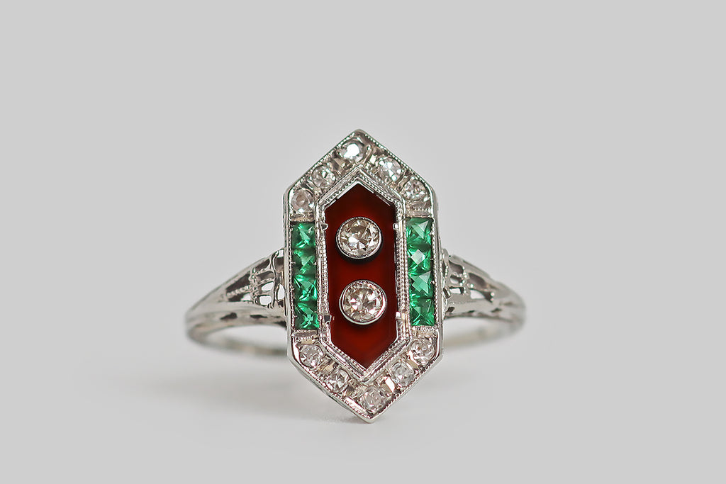 Poor Mouchette | Curated Antique Jewelry, Vintage Jewelry & Engagement Rings | Portland, Oregon | A wonderful, unusual, Art Deco era ring, modeled in 18k white gold, with a long, hexagonal face, cutwork shoulders, and an ornate floral gallery! This 1920s darling is set with a central, polished, carnelian slab, upon whose earthy-red field a pair of small, old European cut diamonds are mounted in bezels. Two groupings of four vibrant, calibre-set, French-cut emeralds 