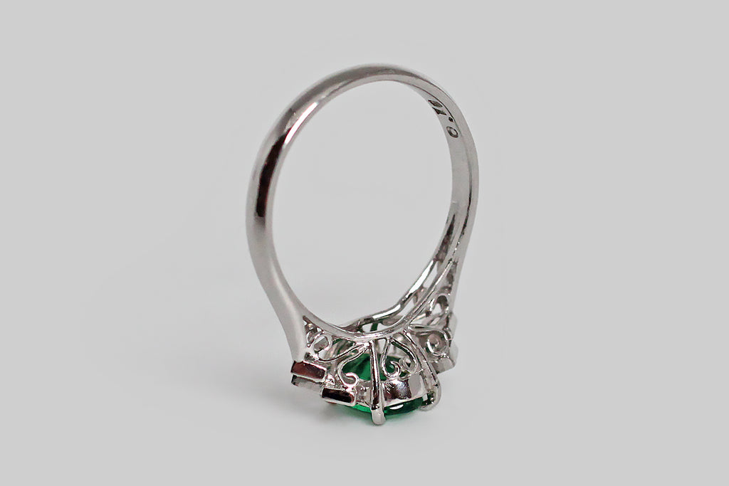 An elegant Art Deco era engagement ring, modeled in platinum, whose primary gem is an especially nice, 3/4 carat, faceted, natural emerald. This vivid central gem (oval) is mounted in a four-prong basket. A pair of decorative, chevron-shaped epaulettes frame the emerald. These shoulder embellishments are really charming— each is composed of three diamond-shaped, diamond-set plaques that sit at staggered heights, coming together to create the chevron shape.