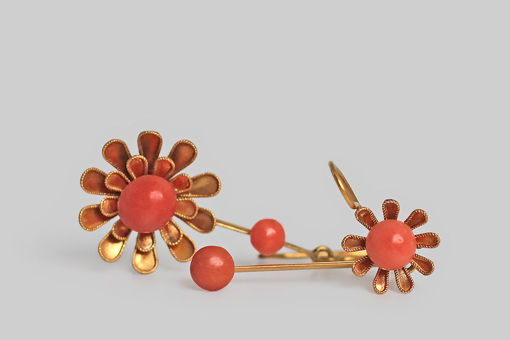 A pair of asymmetrical, coral earrings, fashioned from Victorian-era components. These 18k yellow gold earrings each feature an Etruscan revival (daisy-like) flower— one is larger with two tiers of petals, and one is smaller with a single tier. The flower's petals are edged with delicate, twisted wire, and a round, natural coral cabochon sits at the center of each blossom. 
