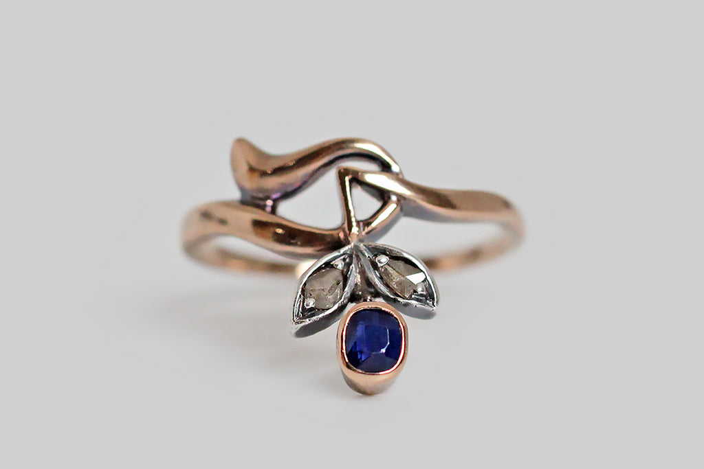 A sweet, Art Nouveau era ring, whose old cut blue sapphire is a flower bud, nodding above a pair of silver-topped, rose cut diamond set leaves. This ring is made in 14k rose gold, with a swooping, asymmetrical band, that curves gracefully and then jags, here and there. Its glinting rose cut diamonds are smoky, and rustic. These small, poetic, Art Nouveau pieces are few and far between. 