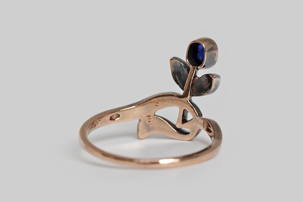 A sweet, Art Nouveau era ring, whose old cut blue sapphire is a flower bud, nodding above a pair of silver-topped, rose cut diamond set leaves. This ring is made in 14k rose gold, with a swooping, asymmetrical band, that curves gracefully and then jags, here and there. Its glinting rose cut diamonds are smoky, and rustic. These small, poetic, Art Nouveau pieces are few and far between. 
