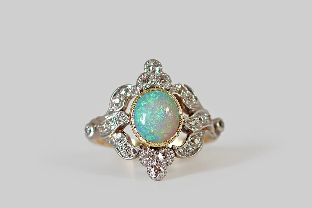 Antique Jewelry Portland, Vintage Jewelry Portland , Antique Engagement Rings | Poor Mouchette | A striking Belle Époque era ring, modeled in 18k yellow gold and platinum, whose primary gem is a beautiful, fiery, Australian opal. This opal is set in a yellow gold bezel, where it hovers inside an ornate, diamond-set, platinum-topped "frame." Sparkling diamond trefoils flank the opal to the north and south, while trios of undulating, ribbon