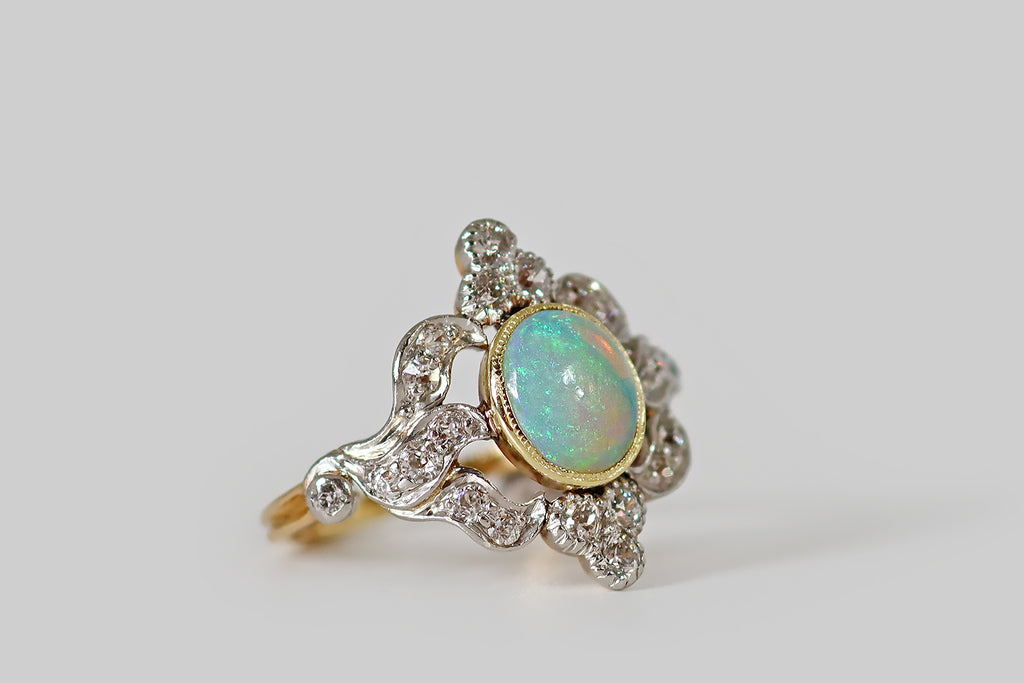 Antique Jewelry Portland, Vintage Jewelry Portland , Antique Engagement Rings | Poor Mouchette | A striking Belle Époque era ring, modeled in 18k yellow gold and platinum, whose primary gem is a beautiful, fiery, Australian opal. This opal is set in a yellow gold bezel, where it hovers inside an ornate, diamond-set, platinum-topped "frame." Sparkling diamond trefoils flank the opal to the north and south, while trios of undulating, ribbon