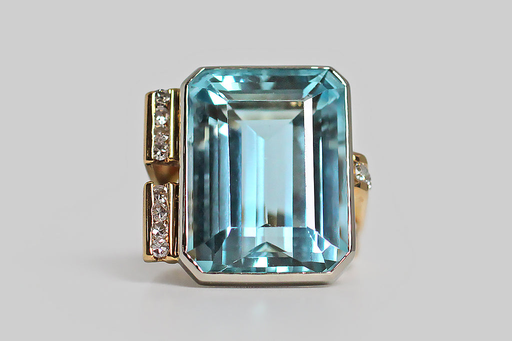 A bold vintage, modernist ring, modeled in 18k gold, featuring a magnificent, twenty-one carat, step-cut aquamarine gem. This vibrant gem is held in a smooth, low-profile, white gold bezel, that perches atop the ring's fully-round shank on four slender stilts. The ring's split shank is modeled in yellow gold, and is fully round. It's shoulders are innovative and asymmetrical— one side features a broad pair of elegant, tapering laths, whose front-facing ends are channel-set with white, brilliant cut diamonds
