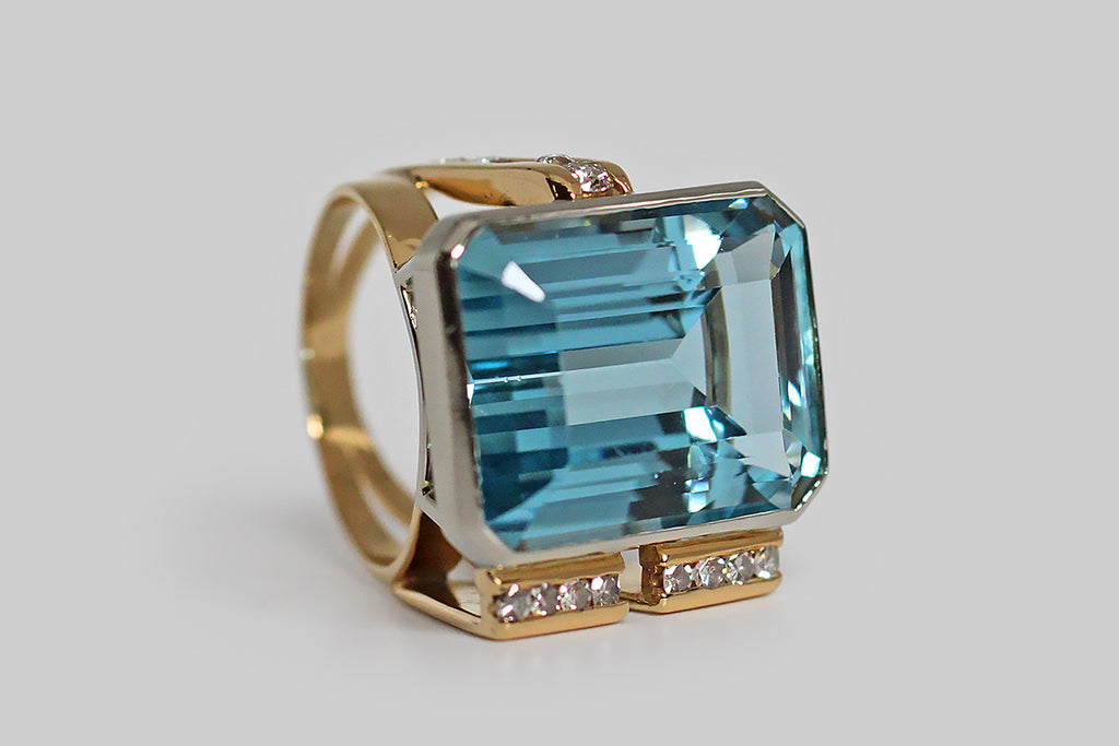 A bold vintage, modernist ring, modeled in 18k gold, featuring a magnificent, twenty-one carat, step-cut aquamarine gem. This vibrant gem is held in a smooth, low-profile, white gold bezel, that perches atop the ring's fully-round shank on four slender stilts. The ring's split shank is modeled in yellow gold, and is fully round. It's shoulders are innovative and asymmetrical— one side features a broad pair of elegant, tapering laths, whose front-facing ends are channel-set with white, brilliant cut diamonds