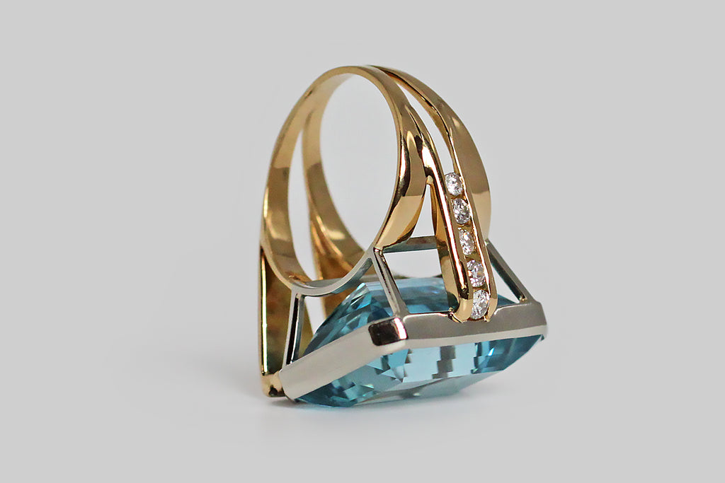 A bold vintage, modernist ring, modeled in 18k gold, featuring a magnificent, twenty-one carat, step-cut aquamarine gem. This vibrant gem is held in a smooth, low-profile, white gold bezel, that perches atop the ring's fully-round shank on four slender stilts. The ring's split shank is modeled in yellow gold, and is fully round. It's shoulders are innovative and asymmetrical— one side features a broad pair of elegant, tapering laths, whose front-facing ends are channel-set with white, brilliant cut diamonds