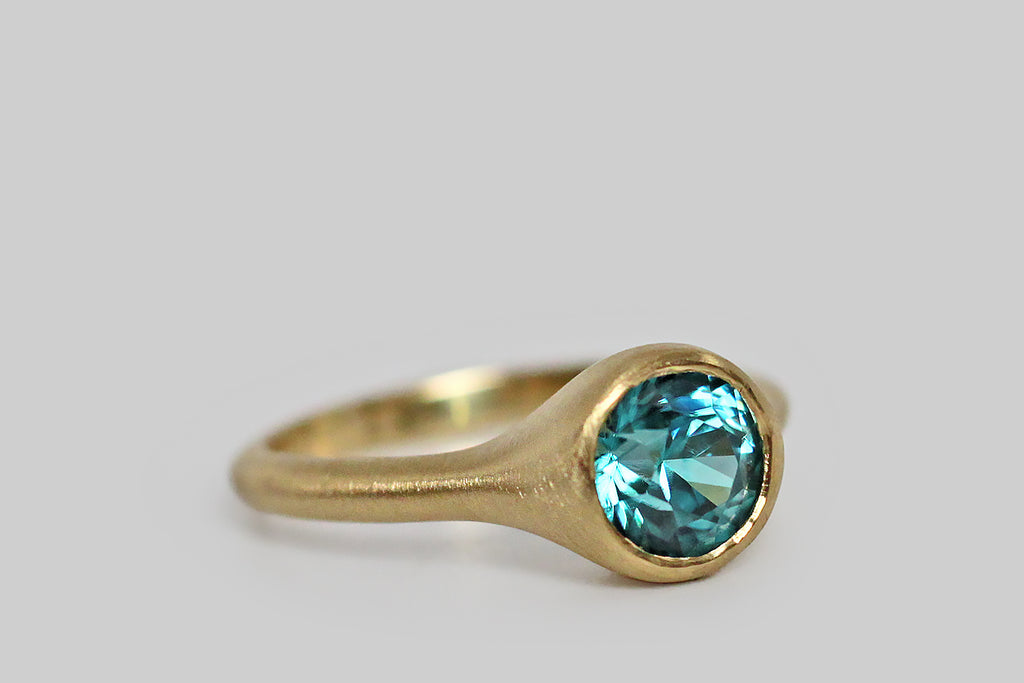 Vibrant and tranquil, a round, faceted, 2.2 carat, blue zircon gem lays nested, inside a thick-walled, organic bezel, which tapers seamlessly into its ring's integral shank. Zircon is an unusual gemstone that has the distinction of being the oldest mineral found on earth, at 4.4 billion years old— in the middle ages it was thought to induce sound sleep, and to drive away evil spirits. This blue zircon gem was originally mined and cut in the 1930s— its facets have been carefully refreshed by a local cutter. 