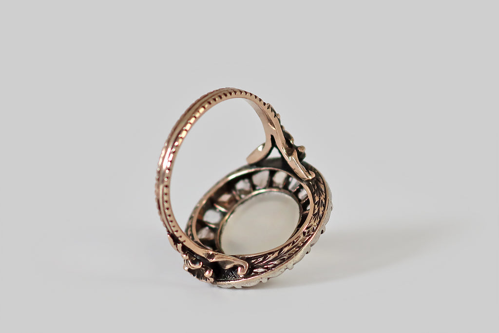 Antique Jewelry Portland, Vintage Jewelry Portland , Antique Engagement Rings | Poor Mouchette | A wonderful, unusual, Edwardian era ring, modeled in rose gold and silver, whose primary gem is a large, round, cat's eye moonstone cabochon. This white moonstone is set in a low-profile, rose gold bezel, where it is surrounded by a halo of eighteen, natural, rose cut diamonds— it has a prominent, glowing, vertical eye that shifts with movement! The old rose cut diamonds