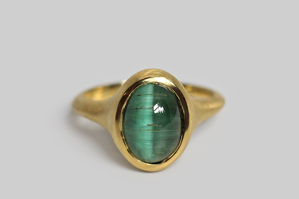 Glowing and vibrant, a highly-saturated, a teal-green tourmaline cabochon sits, nested, inside a thick-walled, organic bezel, which tapers seamlessly into the ring's integral shank. This elegant, oval, tourmaline gem exhibits the rare and beautiful cats eye phenomenon, known as chatoyancy, which gives the stone a two color 