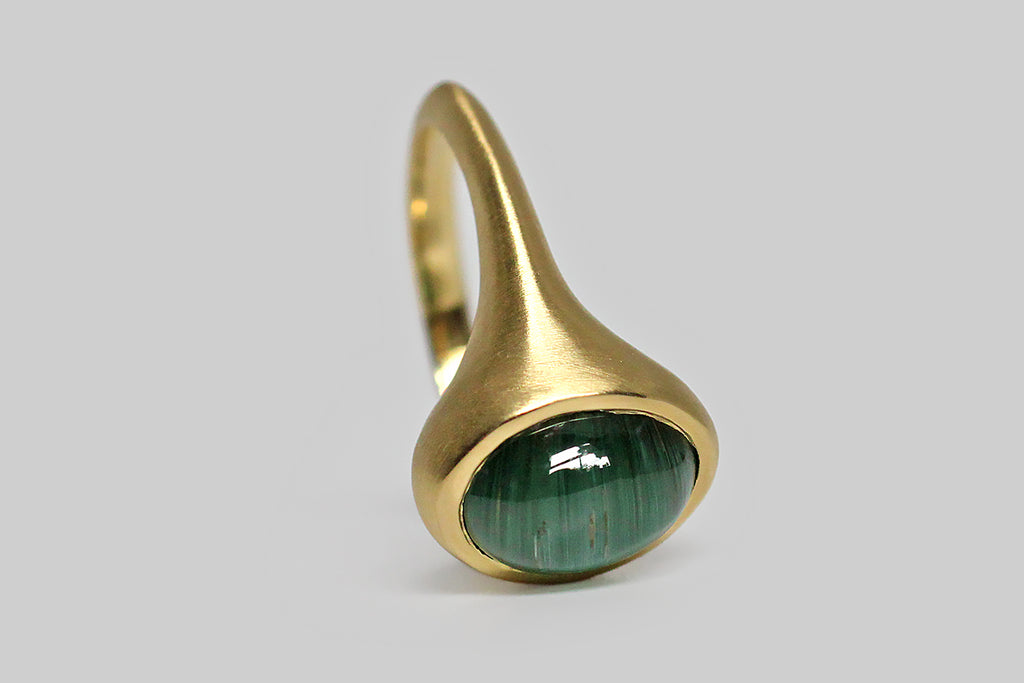 Glowing and vibrant, a highly-saturated, a teal-green tourmaline cabochon sits, nested, inside a thick-walled, organic bezel, which tapers seamlessly into the ring's integral shank. This elegant, oval, tourmaline gem exhibits the rare and beautiful cats eye phenomenon, known as chatoyancy, which gives the stone a two color 