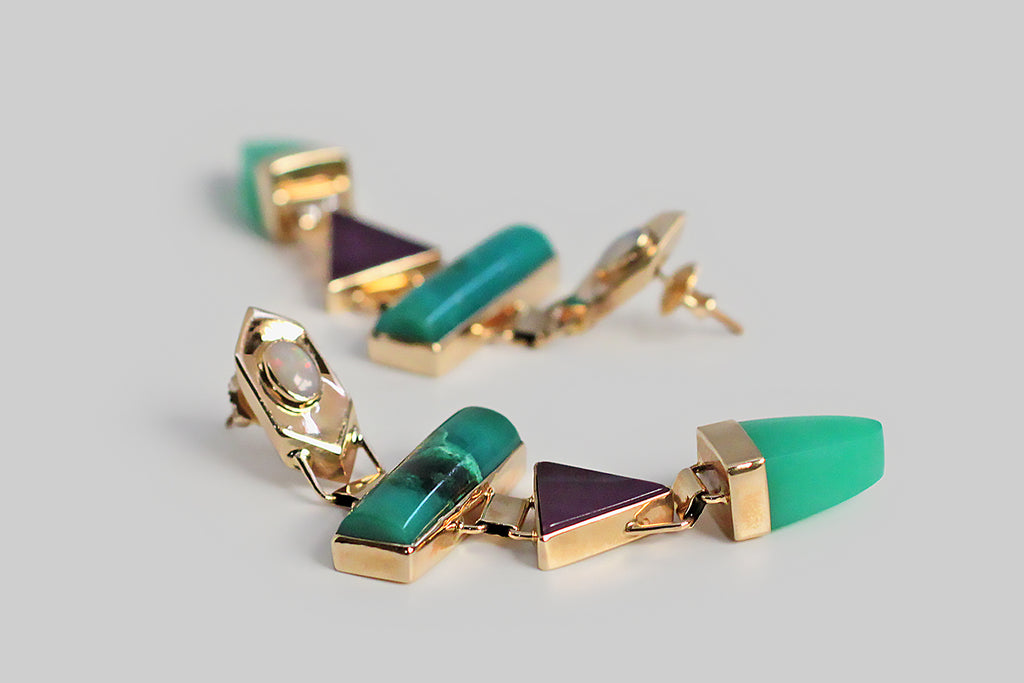 A pair of long, late-vintage drop earrings, modeled in 14k yellow gold, whose playful form concerns itself with shape and color. These earrings feature four types of colorful, bezel-set, cabochon gemstones — white opal, chrysocolla, sugilite, and chrysoprase — these vibrant stones are arranged in a vertical row of chunky geometric shapes. The 14k bezels are beautifully made, and hinged together, so that they swing back and forth with movement. These earrings are entirely hand fabricated. 
