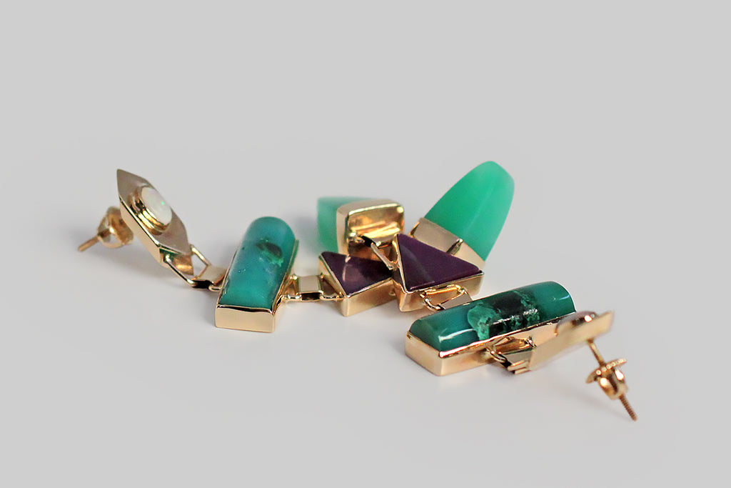 A pair of long, late-vintage drop earrings, modeled in 14k yellow gold, whose playful form concerns itself with shape and color. These earrings feature four types of colorful, bezel-set, cabochon gemstones — white opal, chrysocolla, sugilite, and chrysoprase — these vibrant stones are arranged in a vertical row of chunky geometric shapes. The 14k bezels are beautifully made, and hinged together, so that they swing back and forth with movement. These earrings are entirely hand fabricated. 