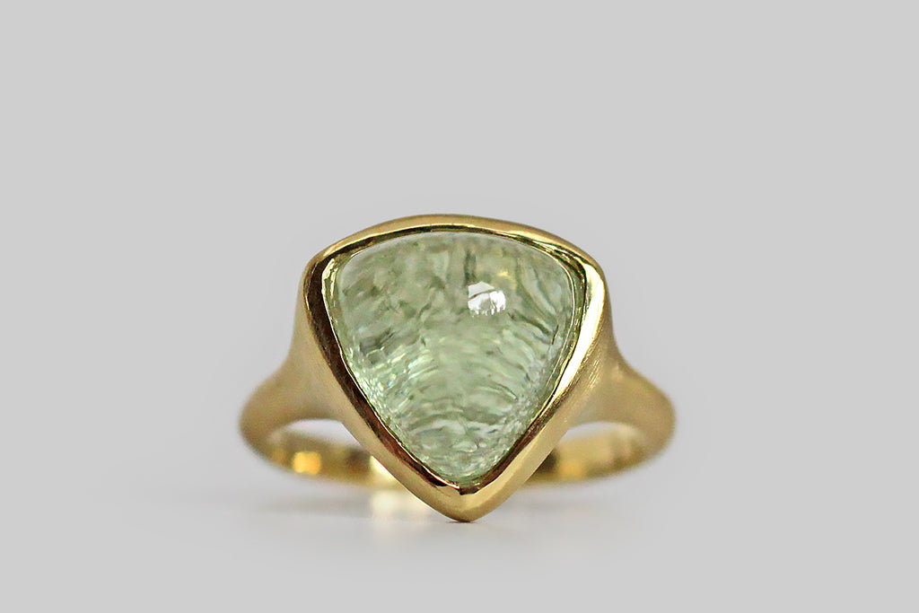 Soothing and glowing, a misty, cucumber-green tourmaline cabochon sits, nested, inside a thick-walled, organic bezel, which tapers seamlessly into its ring's integral shank. This bubbly, triangular, tourmaline gem has been cleverly cut, leaving the natural, crystalline texture of its terminus intact on the bottom side— the gem's smooth high dome magnifies this rough crystal texture to magical effect!