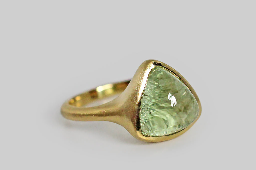 Soothing and glowing, a misty, cucumber-green tourmaline cabochon sits, nested, inside a thick-walled, organic bezel, which tapers seamlessly into its ring's integral shank. This bubbly, triangular, tourmaline gem has been cleverly cut, leaving the natural, crystalline texture of its terminus intact on the bottom side— the gem's smooth high dome magnifies this rough crystal texture to magical effect!