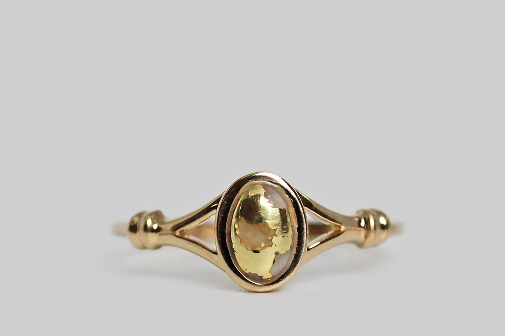 A dainty vintage ring, modeled in 14k yellow gold, featuring a small, oval, gold-in-quartz cabochon. This bezel-set, gold-bearing quartz gem is heavily figured with natural gold, presenting as swaths of vibrant yellow that seem to paint its surface. The ring’s thick-walled bezel floats inside its split shoulders. The shoulders are decorated with a simple banded detail, that makes them appear bound together. The ring's shank is substantial and softly square. 
