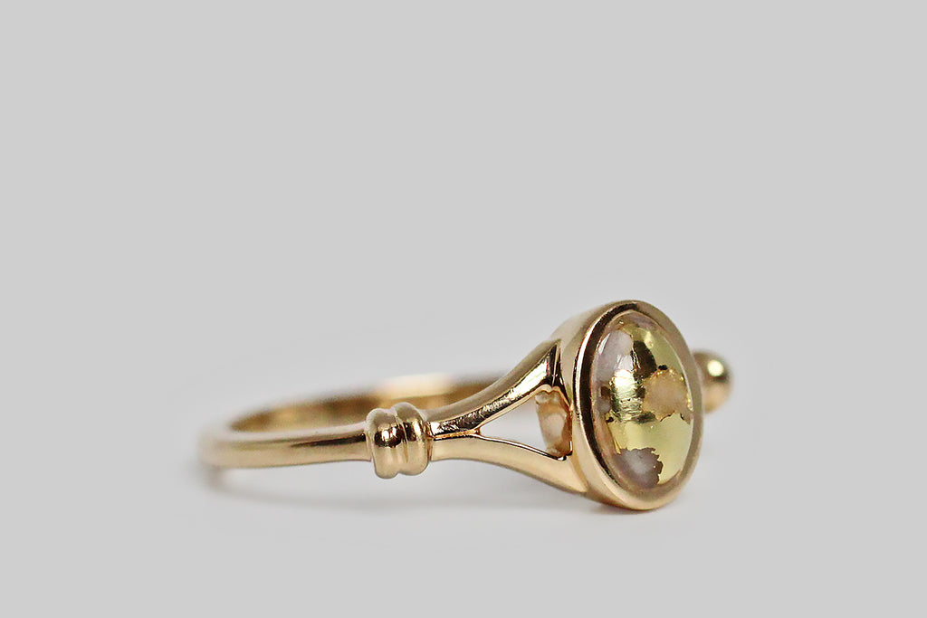 A dainty vintage ring, modeled in 14k yellow gold, featuring a small, oval, gold-in-quartz cabochon. This bezel-set, gold-bearing quartz gem is heavily figured with natural gold, presenting as swaths of vibrant yellow that seem to paint its surface. The ring’s thick-walled bezel floats inside its split shoulders. The shoulders are decorated with a simple banded detail, that makes them appear bound together. The ring's shank is substantial and softly square. 