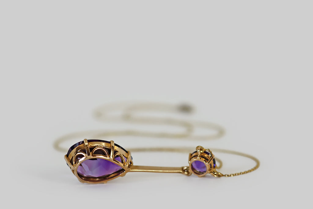 Antique Jewelry Portland, Vintage Jewelry Portland , Antique Engagement Rings | Poor Mouchette | An elegant, Edwardian-era lavalier necklace, modeled in 14k gold, whose focal element is a large, faceted pear shaped amethyst. This lovely faceted pear drops from a single, smaller, amethyst round— a long, slender, knife-edge bar connects the two gems. These old amethysts are a highly-saturated, deep, cool-tone purple.
