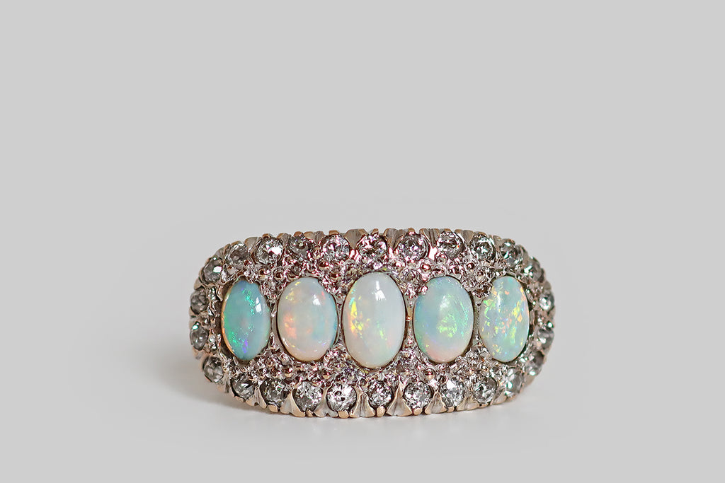 Antique Jewelry Portland, Vintage Jewelry Portland , Antique Engagement Rings | Poor Mouchette | An elegant antique five stone ring, modeled in 14k rose gold, whose primary gems are a series of fiery white opals. These opal cabochons graduate in size across the ring's long, east/west oriented face, which is gently bowed. A border of glittering old cut diamonds, mounted in notched, fishtail settings, surrounds the five opals— the ring's gallery is a decoratively pierced extension of this diamond surround.