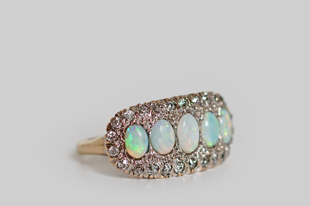An elegant antique five stone ring, modeled in 14k rose gold, whose primary gems are a series of fiery white opals. These opal cabochons graduate in size across the ring's long, east/west oriented face, which is gently bowed. A border of glittering old cut diamonds, mounted in notched, fishtail settings, surrounds the five opals— the ring's gallery is a decoratively pierced extension of this diamond surround.