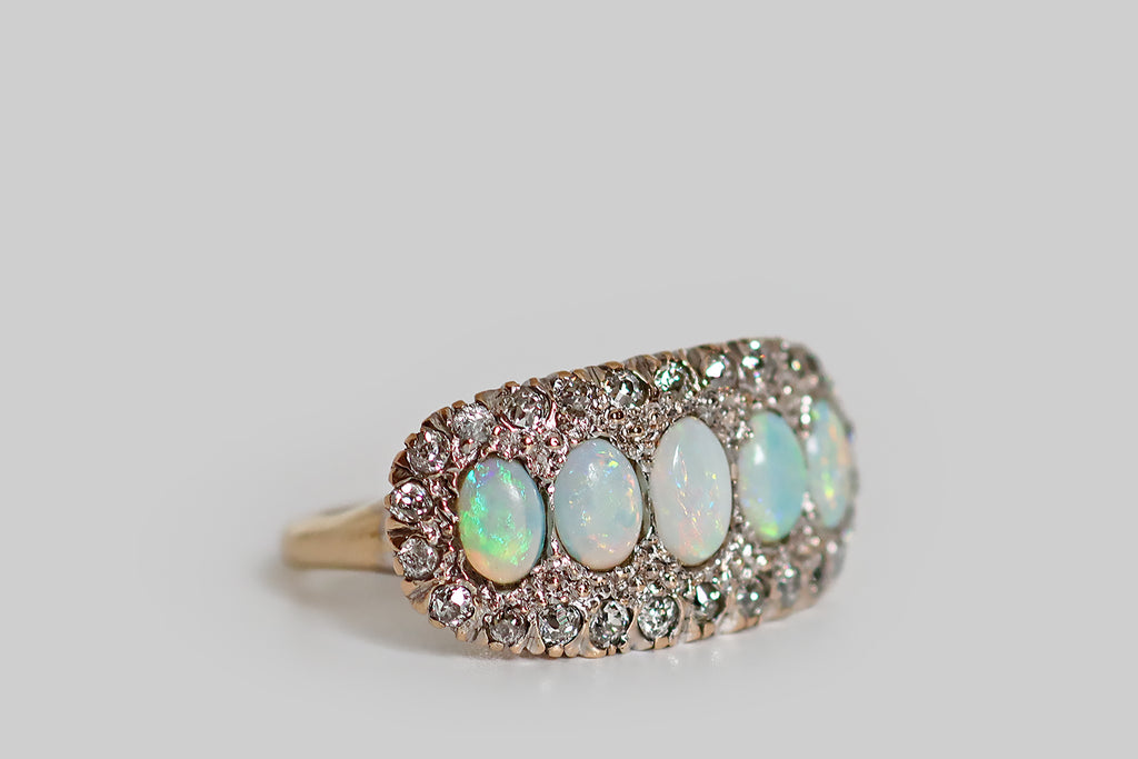 An elegant antique five stone ring, modeled in 14k rose gold, whose primary gems are a series of fiery white opals. These opal cabochons graduate in size across the ring's long, east/west oriented face, which is gently bowed. A border of glittering old cut diamonds, mounted in notched, fishtail settings, surrounds the five opals— the ring's gallery is a decoratively pierced extension of this diamond surround.